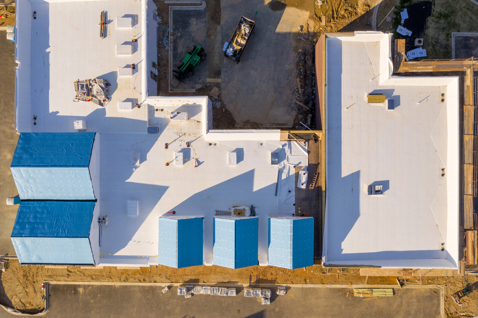 Aerial view of a construction site with white rooftops and blue-roofed sections, surrounded by construction materials, equipment, and dirt areas.