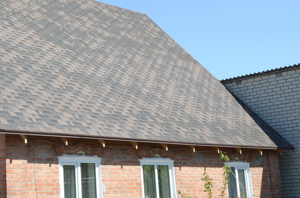 A house with a steeply sloped roof covered in gray asphalt shingles, red brick walls, and three white-framed windows under clear blue sky.