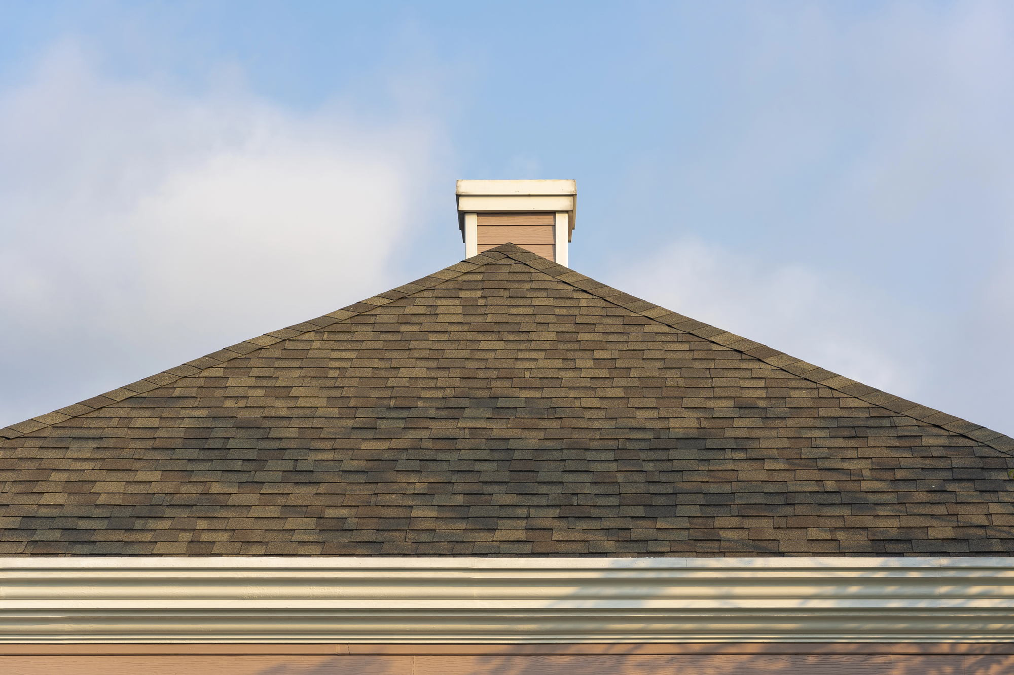 A close-up view of a shingled, triangular roof with a small chimney against a partly cloudy sky.