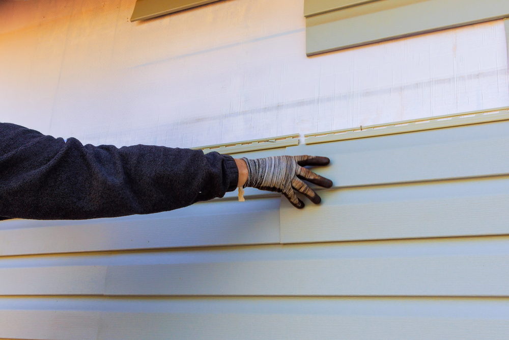 A person wearing a glove installs light blue vinyl siding panels on the exterior wall of a building.