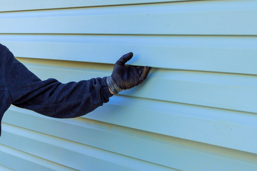 A person wearing a black glove and dark sweatshirt inspects light blue vinyl siding on a building.