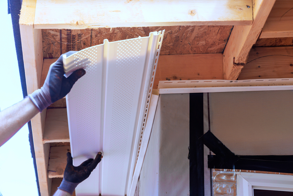 A person wearing gloves installs a white perforated vinyl soffit panel underneath a wooden roof overhang of a house under construction.